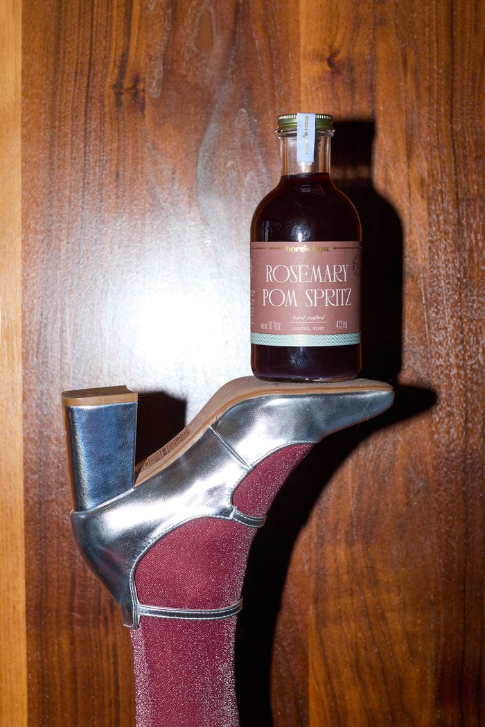 Rosemary Pom Spritz Cocktail Mixer bottle by Here's How, perched on a silver high heel with a wooden backdrop.
