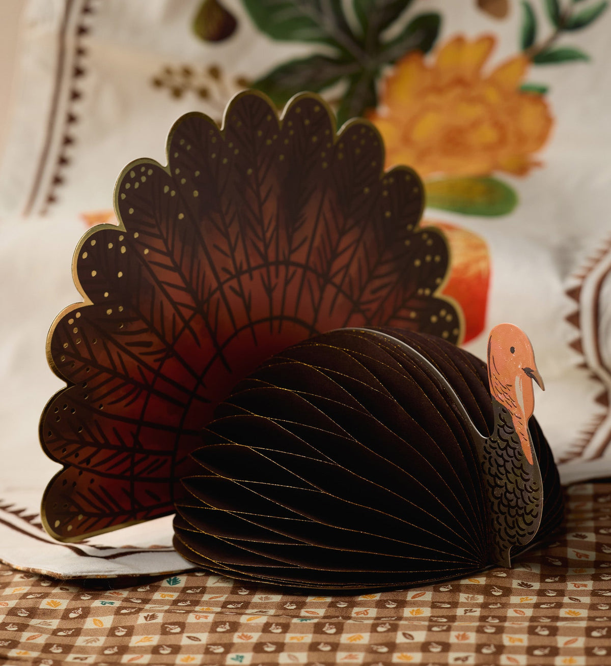 Rifle Paper Co. Turkey Honeycomb Decor on a patterned tablecloth, with fanned brown and orange tail, perfect for Thanksgiving.