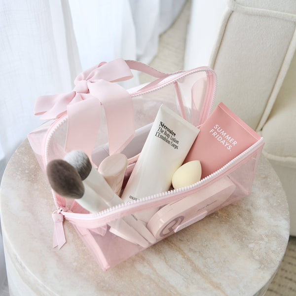 Case-Mate Jelly Makeup Bag, clear pink with a ribbon, filled with beauty products like lotion and brushes on a table.