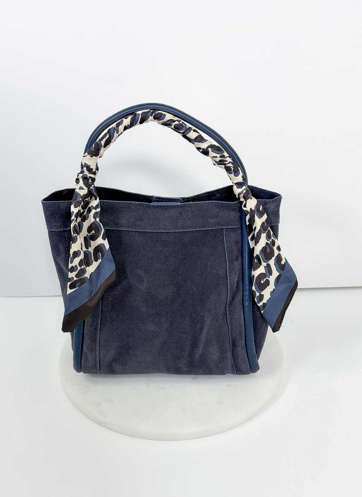 Gianna Suede Bag by BC Bags, a navy blue suede handbag with a leopard print scarf tied to one handle, on a white base.