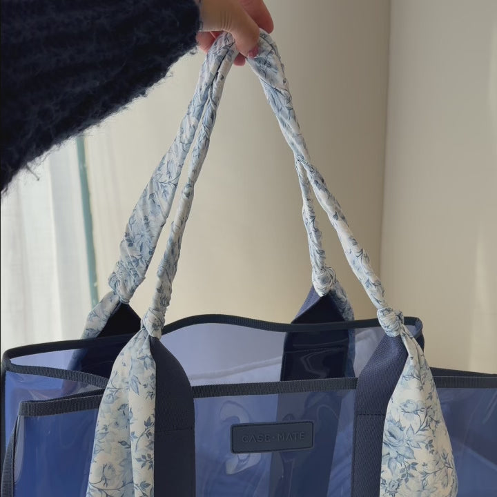 Case-Mate Jelly Stadium Bag Something Navy Twilly. A hand holds a transparent blue tote bag with floral scarf-wrapped handles.