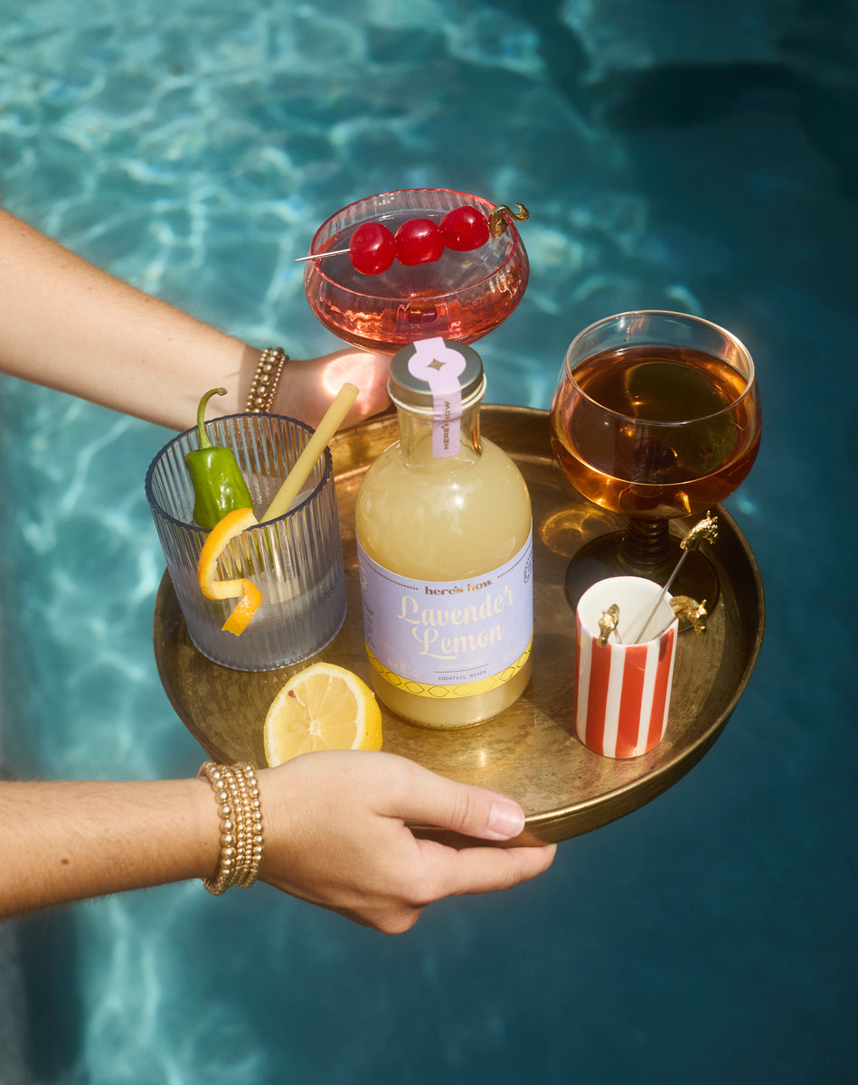 Here's How Lavender Lemon Cocktail Mixer on a tray with cocktails, garnishes, and lemon, held over a sparkling pool.